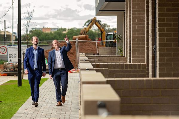 Two men in blue business suits and high visibility vests and white hard hats stand in front of a fenced off, newly developed property and point at vacant space.